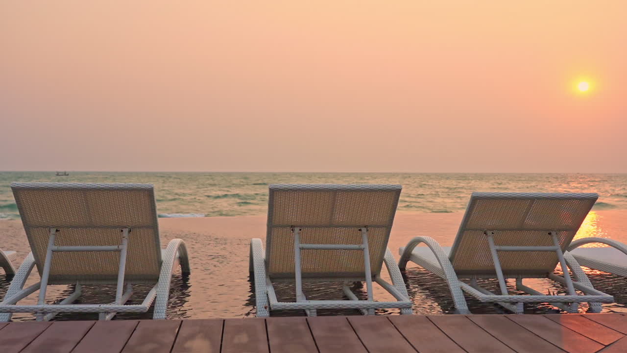 Line of deck chairs on a tropical beach with a beautiful colorful sunset. Seaside lounge with infinity pool and chaise-longues in a pool