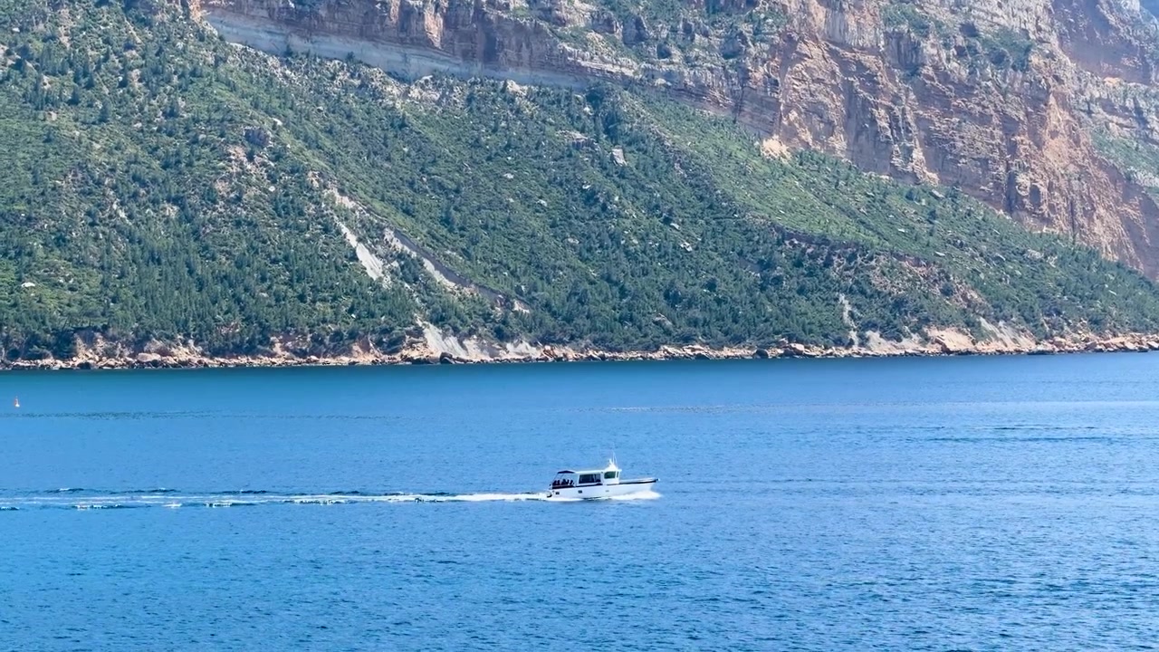 Un motoscafo bianco naviga su acque calme e blu sotto imponenti scogliere calcaree alla luce del sole.
