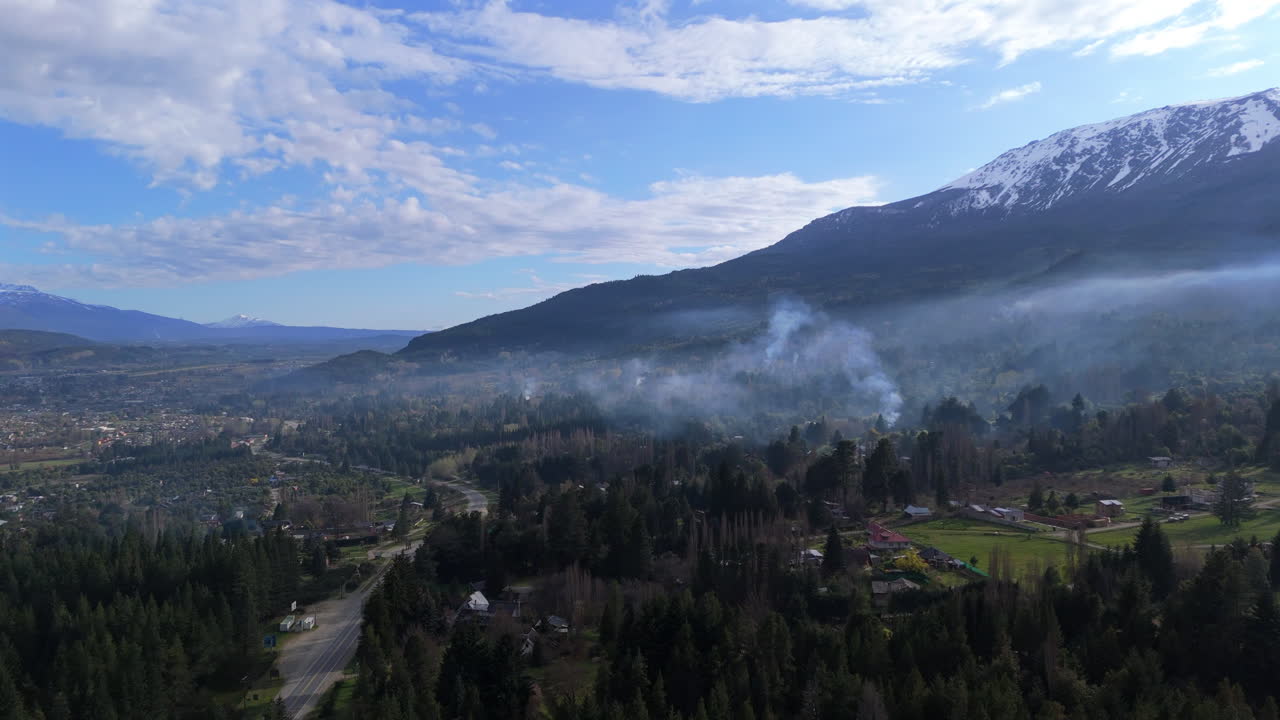 Fast parallax wide angle drone shot over El Bolsón town in Argentinean South with big trees, houses, buildings, some smoke and the Andean mountain range in background. Shot on DJI Air 3 at 4K-60fps.
