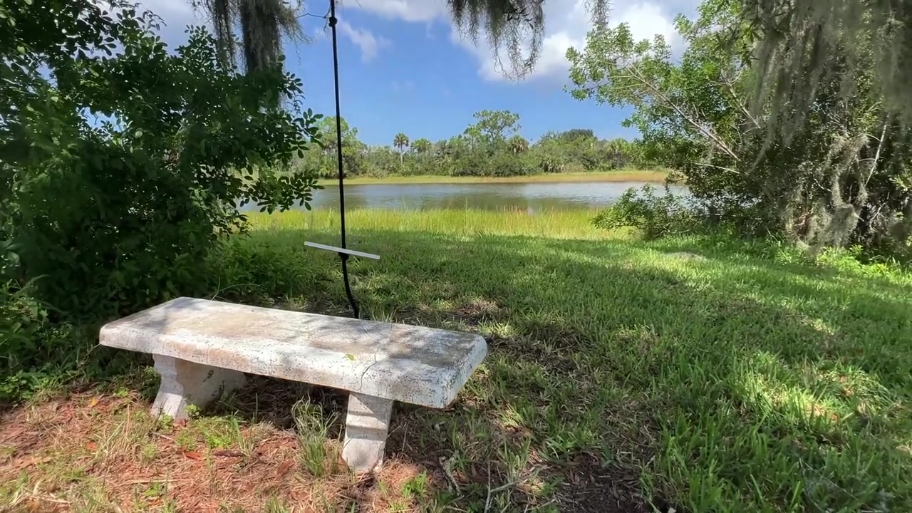 Un banco de piedra desgastado con vistas a un tranquilo lago de Florida rodeado de césped verde, árboles maduros y musgo español colgante bajo un cielo azul brillante