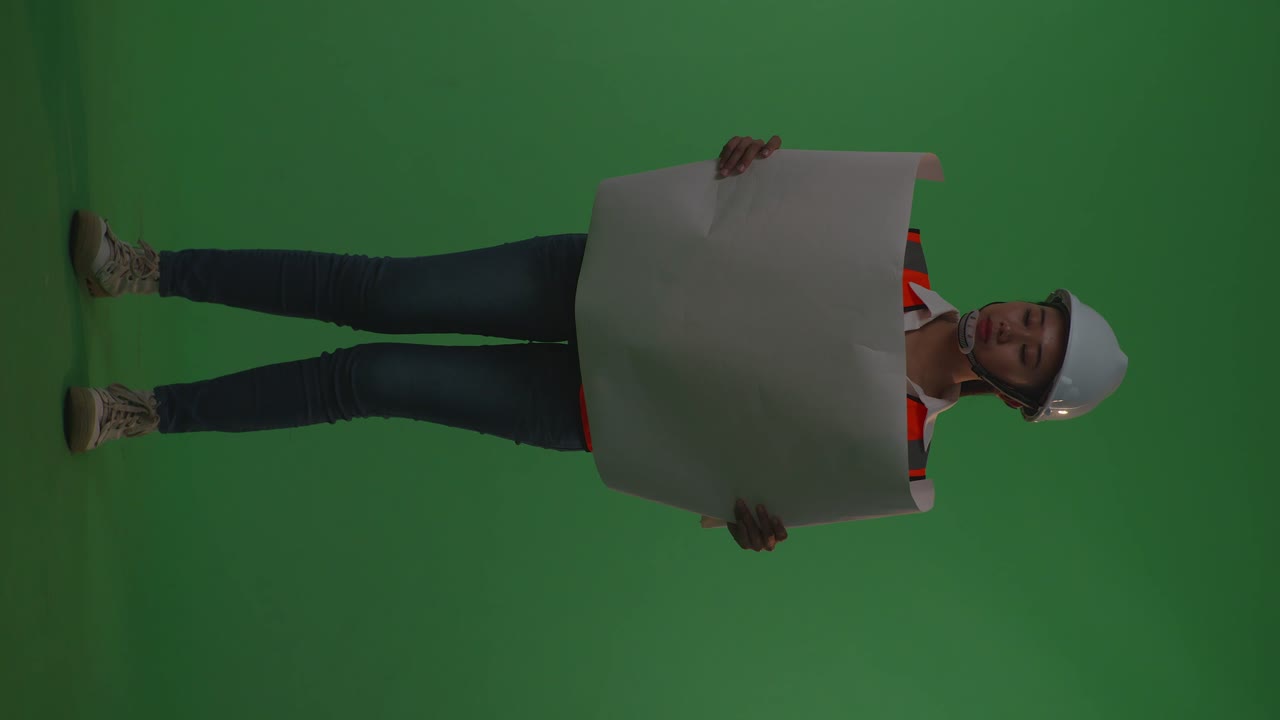 cuerpo lleno de ingeniera asiática con casco de seguridad mirando el plano en sus manos mientras está de pie en el estudio de fondo de pantalla verde, durante la puesta o el amanecer