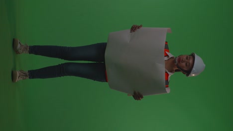 full body of asian female engineer with safety helmet looking at blueprint in her hands while standing in the green screen background studio, during sunset or sunrise time