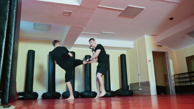 Atleti che si allenano in un'accademia di arti marziali, indossando guanti e abiti neri, praticando tecniche di sport da combattimento con pugni e movimenti difensivi su un pavimento in tatami rosso circondato da sacchi da boxe
