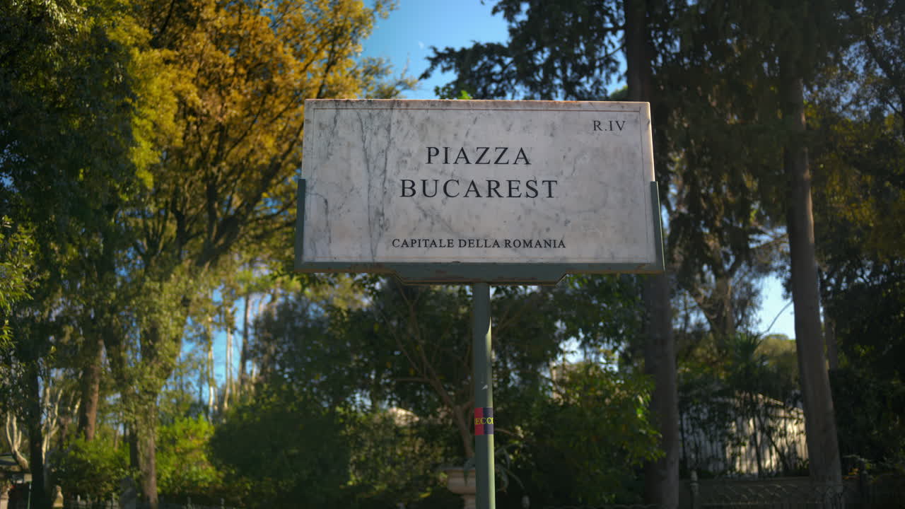 Marble sign of Bucharest square surrounded by trees in sunlight. Rome, Italy