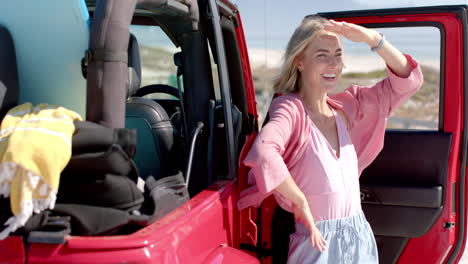 young caucasian woman shields her eyes from the sun beside a red car on a road trip