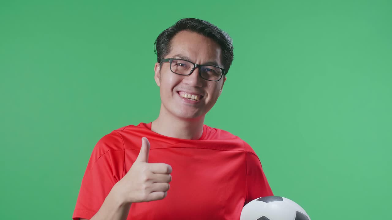 primer plano de un hombre asiático con una pelota mostrando los pulgares hacia arriba mientras anima el fútbol en el fondo de la pantalla verde