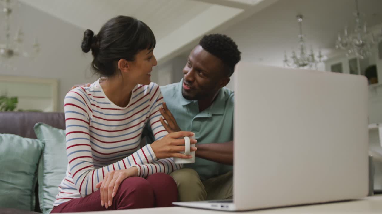 Happy diverse couple sitting on couch and using laptop in living room