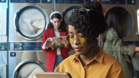 pretty and happy girl in yellow glasses watching funny video on tablet device, resting and waiting for clothes to be washed