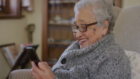 retrato de una mujer mayor alegre usando un teléfono inteligente enviando mensajes de texto navegando disfrutando de la comunicación por teléfono móvil sonriendo enviando mensajes de texto en la sala de estar de la casa usando gafas