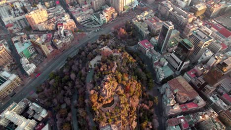 aerial dolly in of santa lucia hill covered in autumnal trees, traffic in alameda avenue and santiago buildings in back, chile