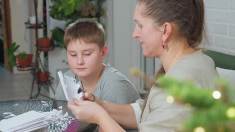 close view of woman carving paper ornament with scissors while son in gray shirt watches attentively, sitting together by table covered with white papers, cozy home setting with potted plants visible