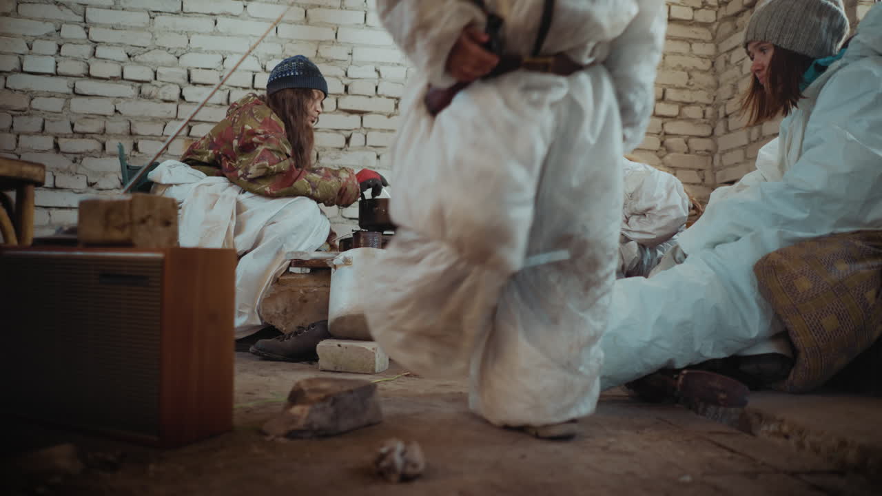 Survivors in protective clothes and winter hats sit inside ruined brick shelter as leader cooks meal on old stove while others wait for food, little boy gets up and walk away