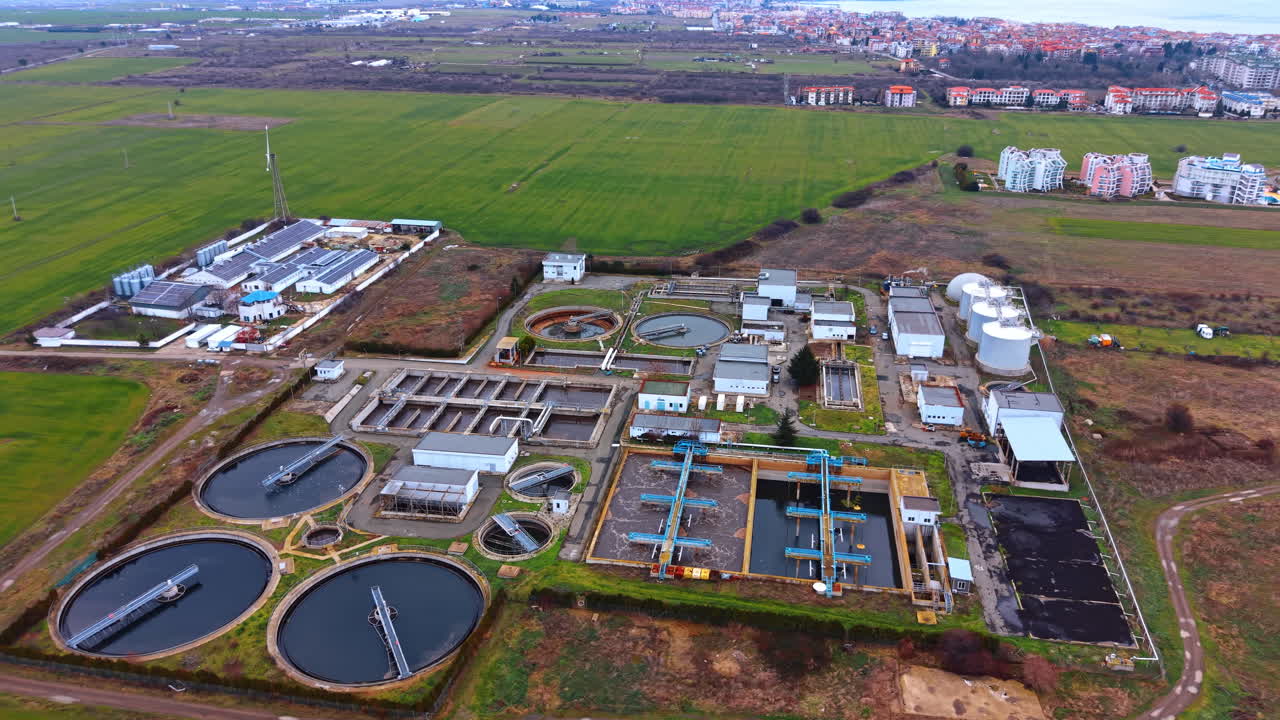 Aerial view of wastewater treatment plant and industrial farm. High-angle wide shot of circular and rectangular water purification tanks next to buildings with solar panels