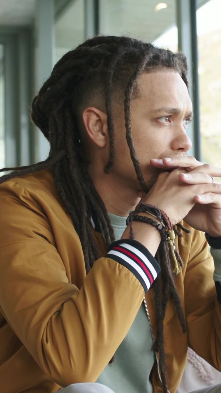 Video verticale: Uomo seduto con i dreadlocks, che guarda fuori dalla finestra di un caffè, in giacca, con fasce, spazio per il testo.