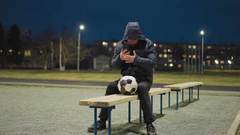 a man is sitting alone on a bench in an empty stadium at night, with a soccer ball between his legs, he reaches into his jacket to retrieve his phone to answer a call