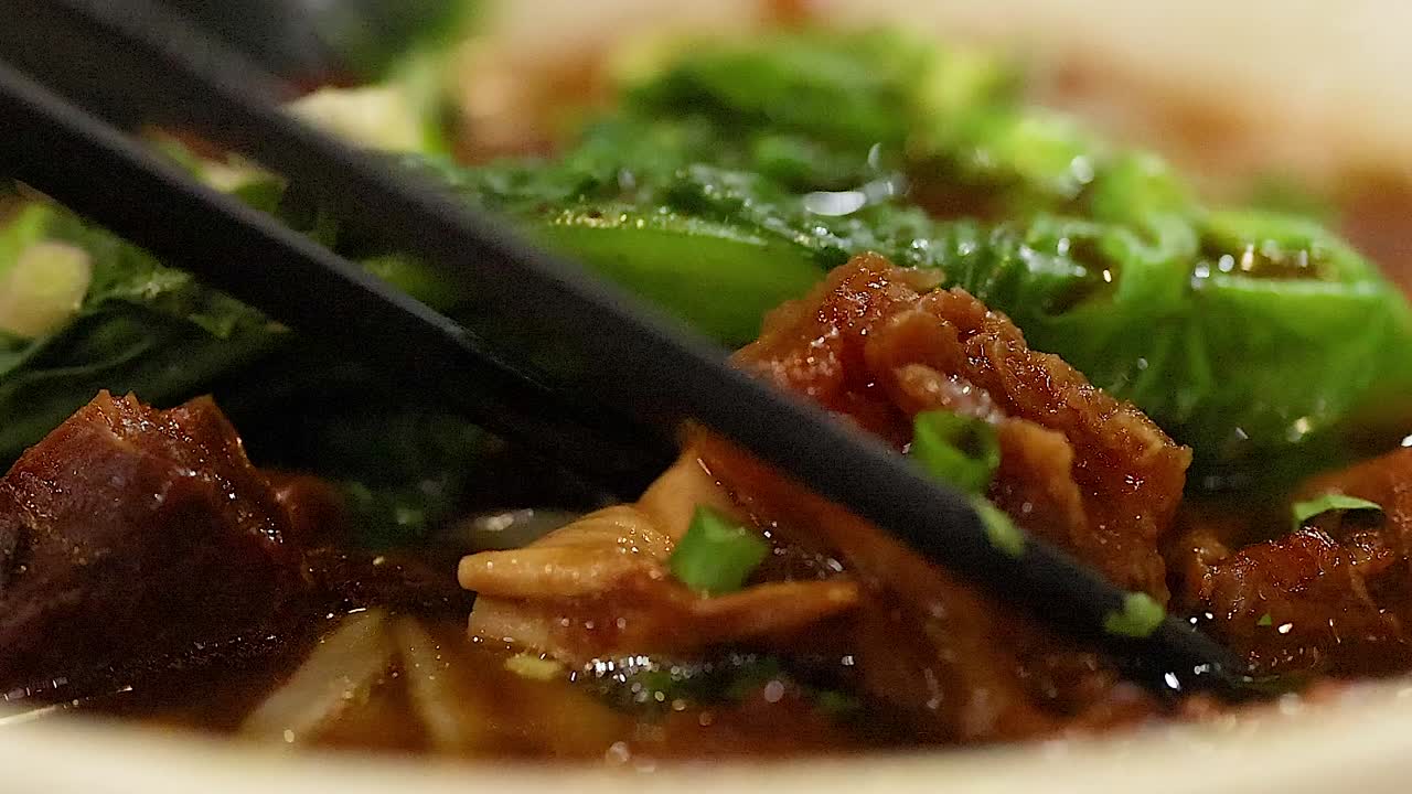 Close-up of chopsticks lifting tender meat from a vibrant noodle bowl with greens.