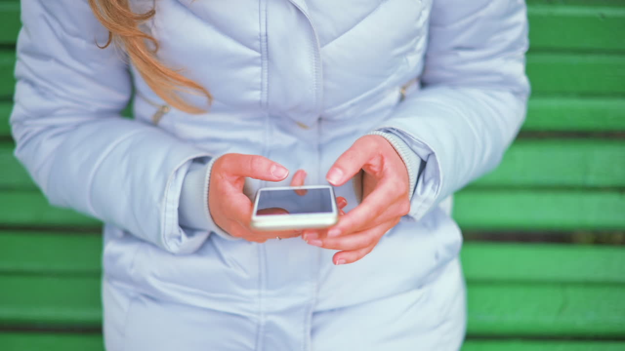 Donna con giacca blu che scorre il telefono seduta su una panchina verde nel parco