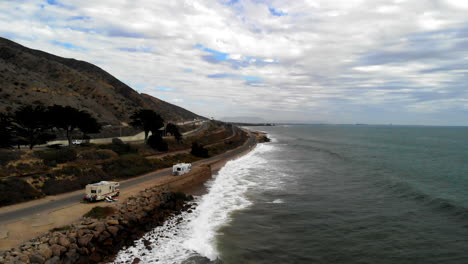 Aerial-drone-shot-low-over-the-ocean-waves-next-to-RV-campers-and-the-cliffs-along-the-101-freeway-in-Ventura,-California