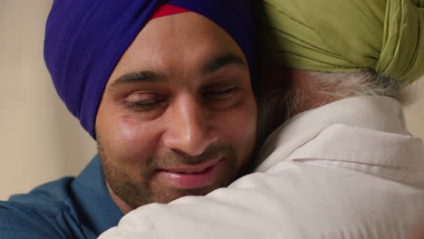 close up studio shot of adult sikh son embracing senior father both wearing turbans against plain background