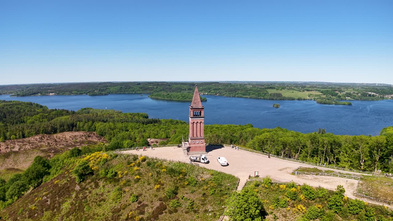 A drone soars high above the Himmelbjerget tower perched on a peninsula, revealing a panoramic view of the surrounding lake and lush green forest in Denmark