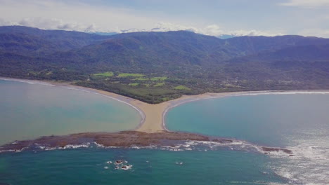 fotografía aérea que se aleja de la ballena en forma de cola punto rocoso de punta uvita rodeado de brillantes olas azules del océano y montañas en la distancia en el sur de puntarenas, costa rica