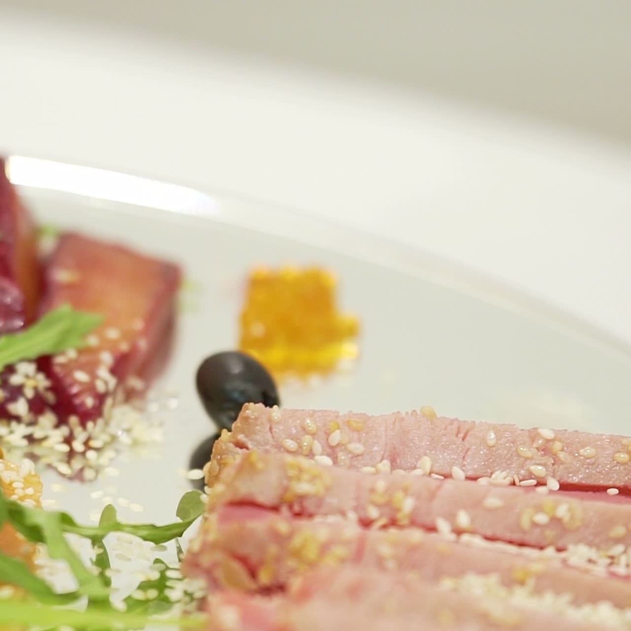 Sliced bacon with sesame is decorated with cherry tomato near pieces of dry-cured meat and lemon with olives and cilantro on a transparent plate on the blurred background in the cafe.