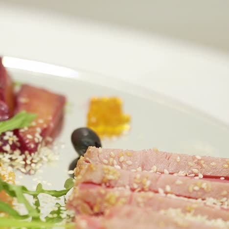 sliced bacon with sesame is decorated with cherry tomato near pieces of dry-cured meat and lemon with olives and cilantro on a transparent plate on the blurred background in the cafe.