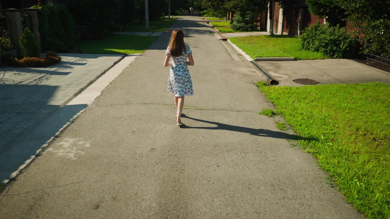 Rear view of lady in white floral dress strolling along quiet residential street on sunny day, gentle breeze blowing her skirt as she walks calmly past houses and greenery with shadow trailing behind