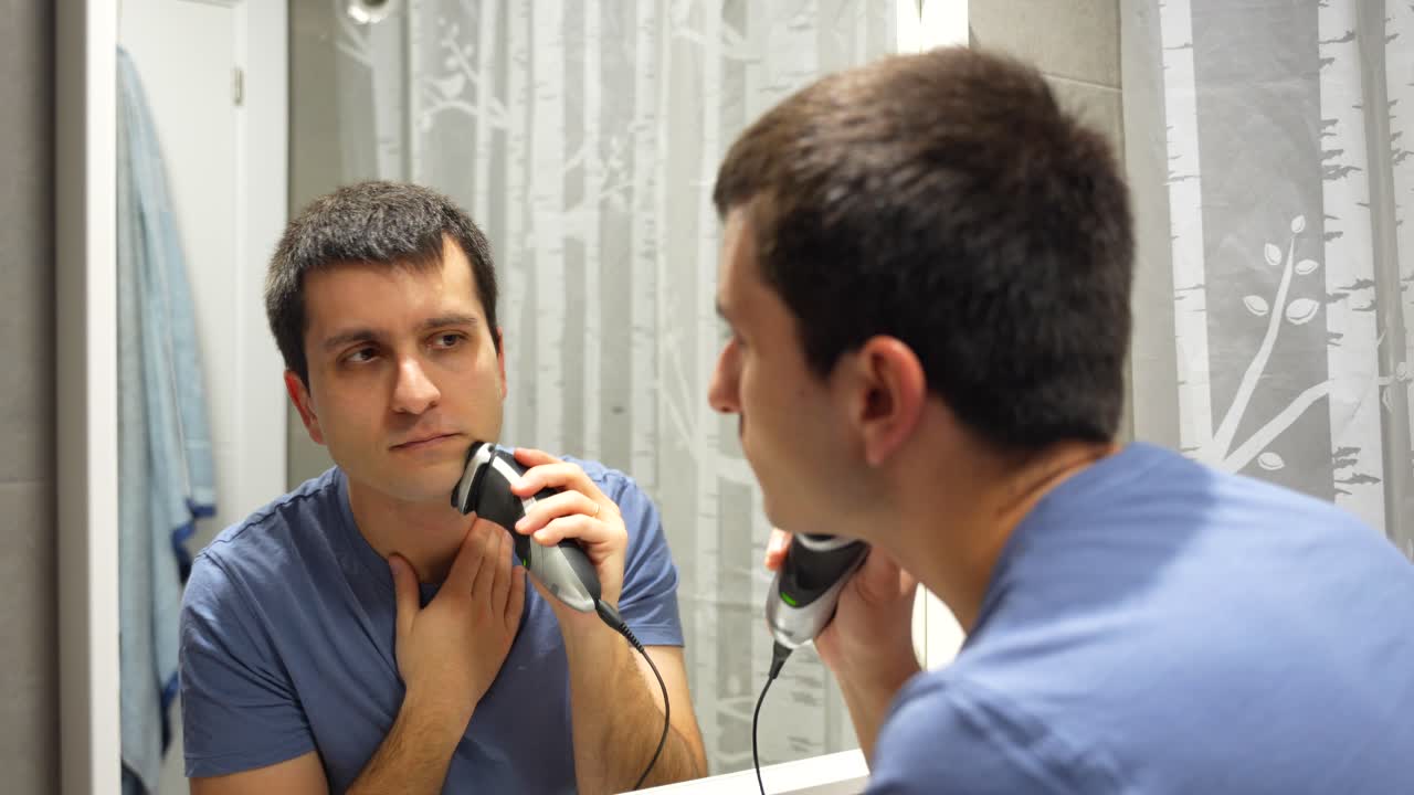 A man shaves his face with an electric shaver in a bathroom mirror