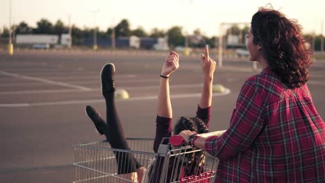 back view of a young woman pushing a grocery cart with her girlfriend inside in the parking by the shopping mall during sunset