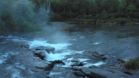 aerial drone shot over a dark misty stream and small waterfalls at sunset with forest trees in silhouette