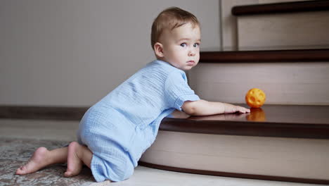 nice cute child kneeled near the stairs indoors and turned his head to look back. lovely boy baby boy pushes the ball from the stairs.