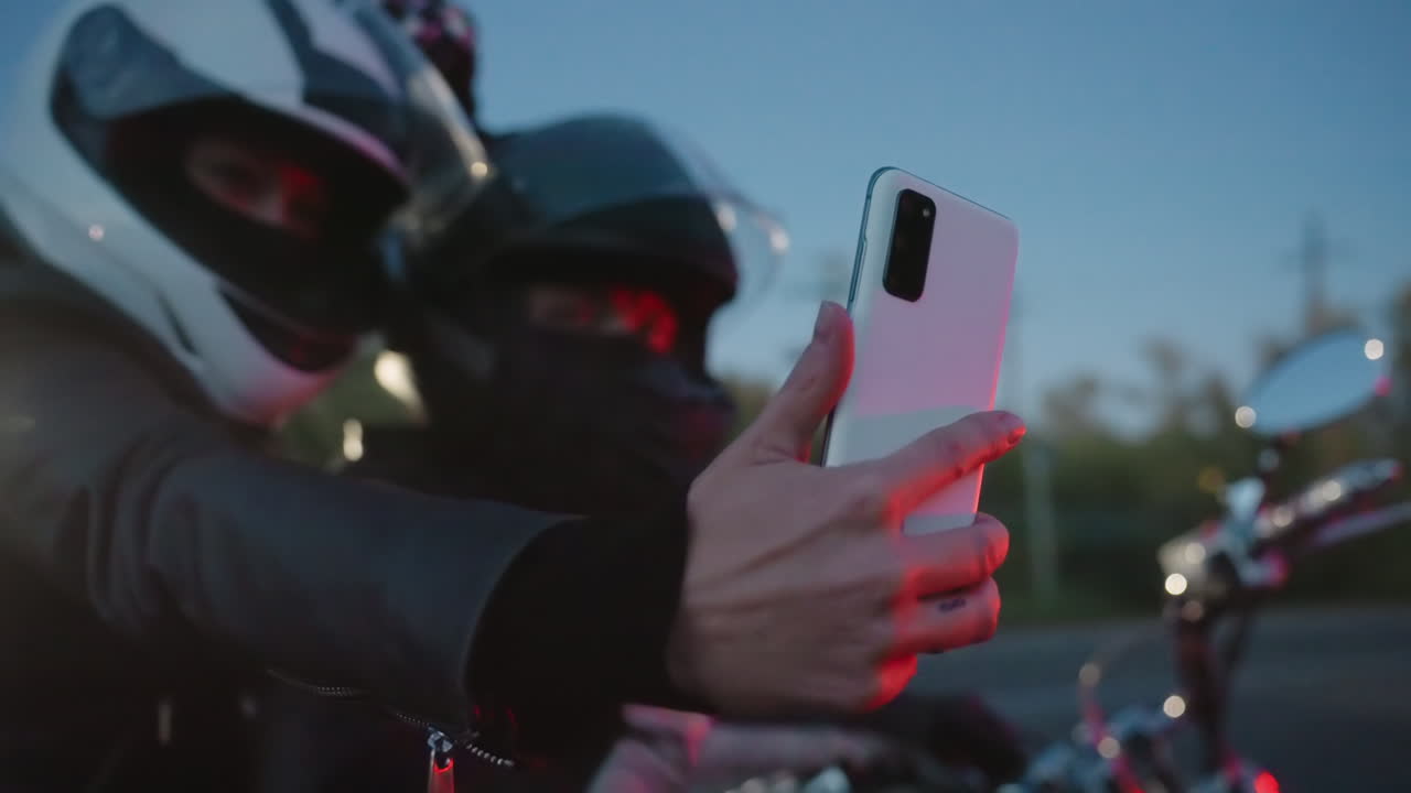 Girlfriend wearing helmet takes selfie with boyfriend while sitting on motorcycle during evening ride, hand stretched forward holding phone as dim street light creates dramatic glow on background