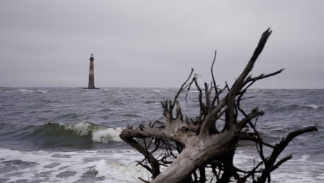 El-Faro-De-La-Isla-Morris-Frente-A-La-Costa-De-Carolina-Del-Sur-En-Segundo-Plano-Mientras-Las-Olas-Chocan-En-Primer-Plano