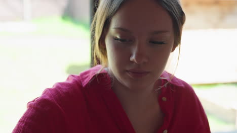 young woman in red top sits near bright window, gently rubbing palms and licking hand in quiet thoughtful moment, with wine glass containing red drink resting on table in soft natural light