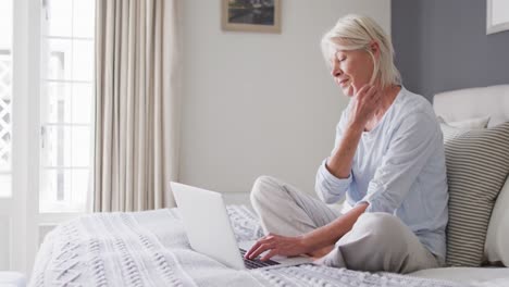 Happy-senior-caucasian-woman-sitting-on-bed-in-bedroom,-using-laptop