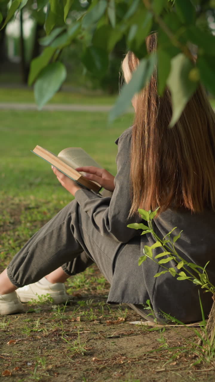 vista trasera de una dama sentada bajo un árbol leyendo un libro, mano en la página, apoyada contra el tronco, la luz del sol ilumina suavemente su cabello y espalda, las piernas ligeramente estiradas, rodeada de vegetación