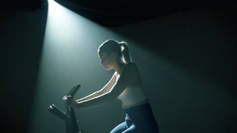 Profile view from knees up of a woman training on a bike in a dark studio. Backlit dramatically, the camera rushes in toward her concentrated, sweaty face, capturing intensity and cinematic tension