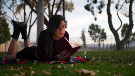 una mujer adulta joven leyendo una novela o un libro de cuentos en el parque al atardecer tumbada en la hierba con hojas de otoño en cámara lenta