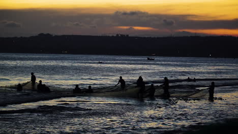 silhouette of group of fishermen retrieving huge fishing net from beach shore