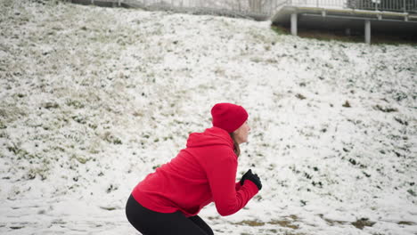 vista lateral de una mujer realizando saltos en cuclillas durante el entrenamiento de invierno al aire libre en terreno nevado con sudadera roja y guantes negros, la escena incluye colina nevada con riel de hierro, atmósfera de niebla