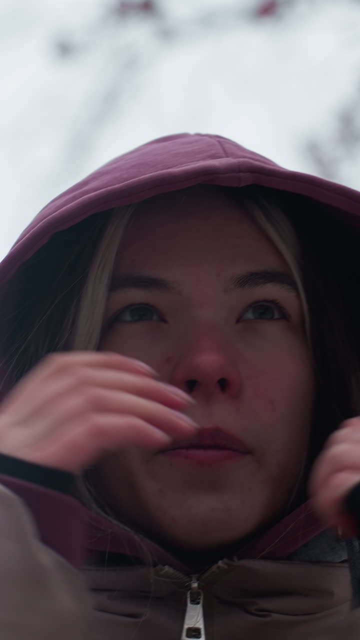 차가운 겨울 날씨에 은 코와 집중된 표정으로 머리 위에 후디를 당긴 젊은 여성, 따뜻함을 위해 후드를 조정하는 동안, 벌거벗은 나무 가지와 함께 흐릿한 얼어붙은 배경