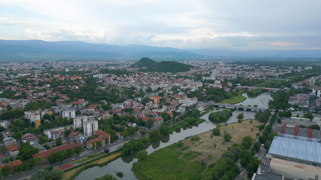 Wide aerial view of Plovdiv, Bulgaria, with the Maritsa River and bridges in the foreground, and distant hills under a cloudy spring sky at sunset