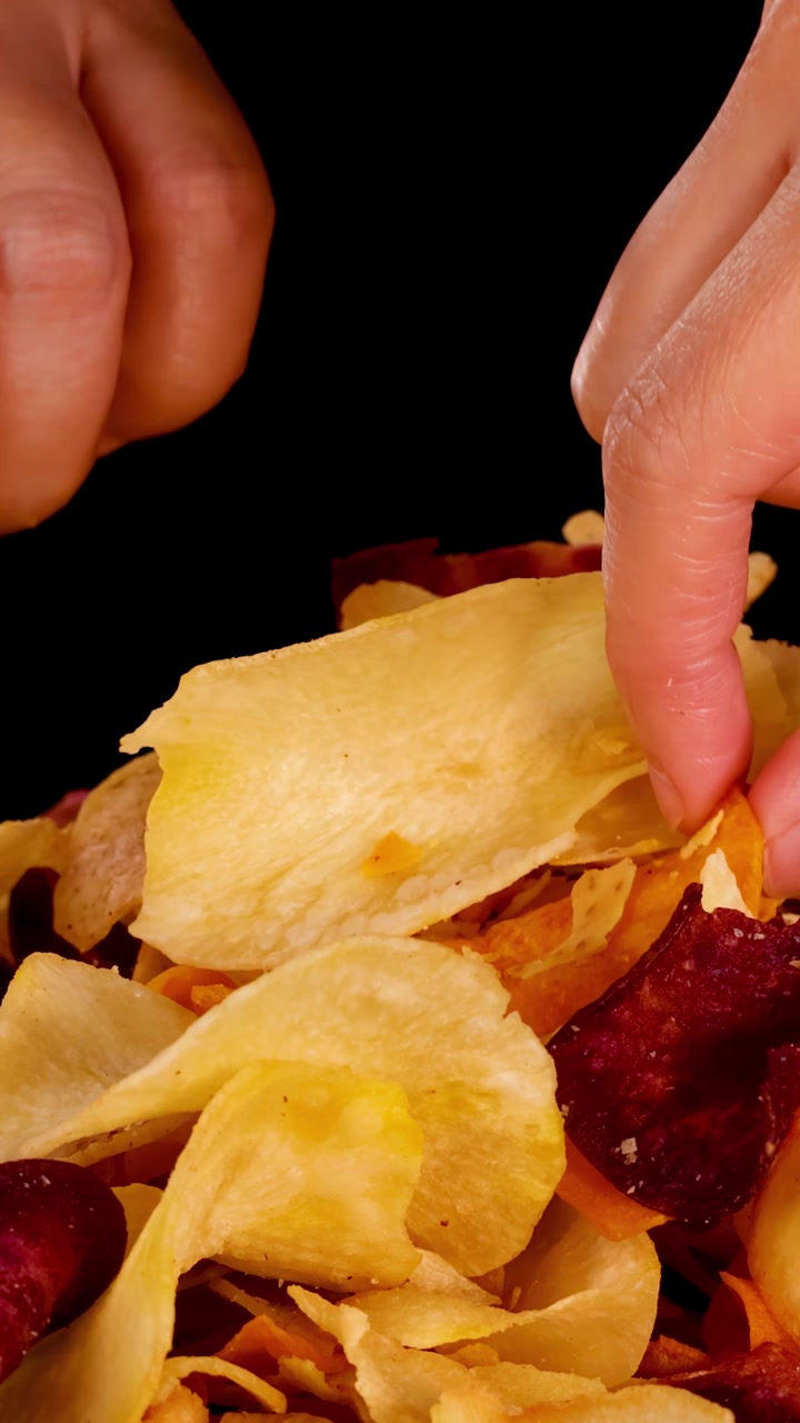 Hand selects crispy mixed root vegetable chips under bright studio lighting, close-up, black background
