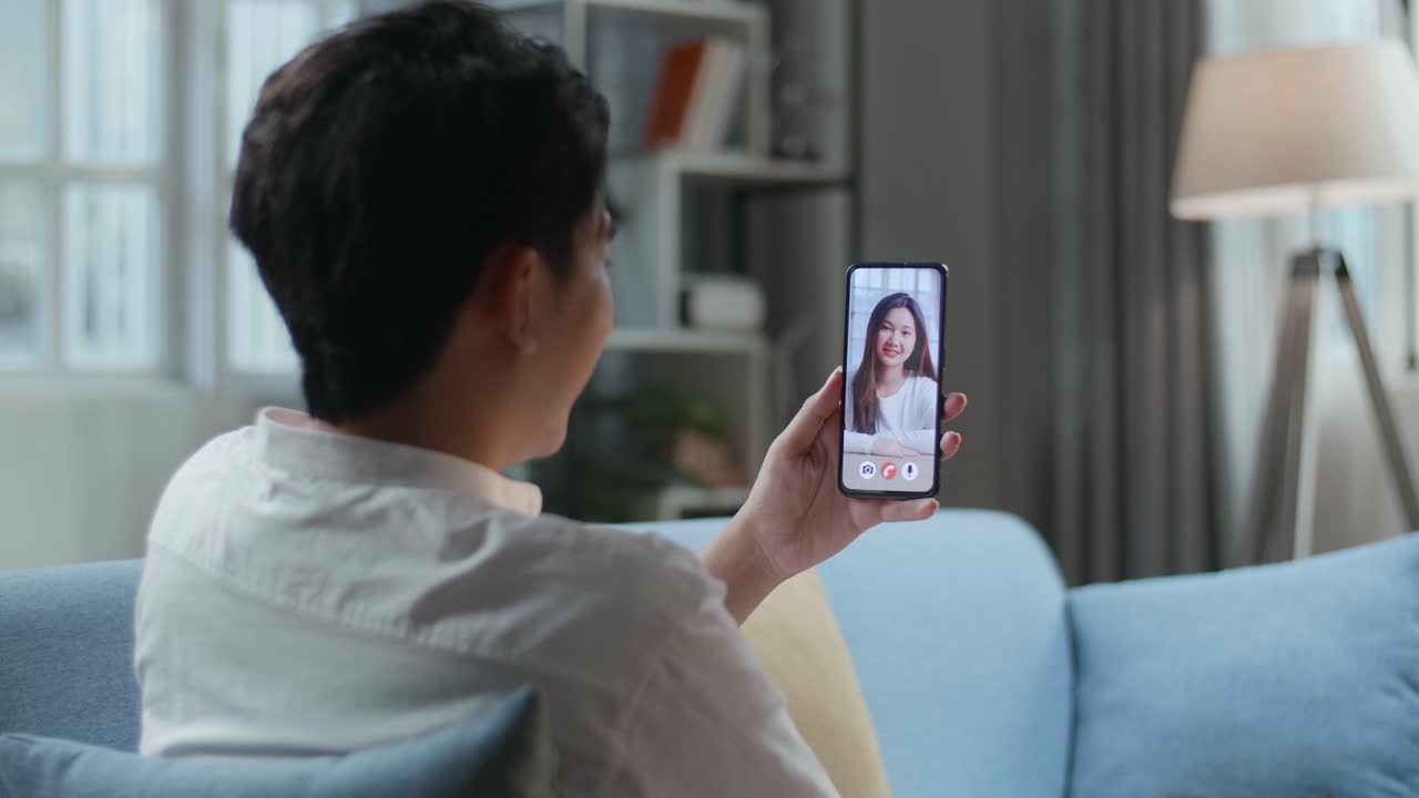 hombre haciendo una llamada de video con una mujer en un teléfono inteligente mientras está tendido en el sofá en la sala de estar