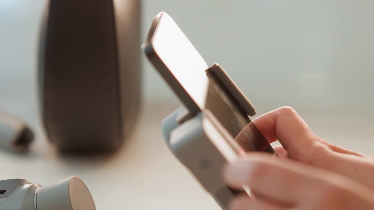 İki elin ince bir akıllı telefonu gimbal montaj plakasına dikkatlice yerleştirdiği yakın çekim; cihaz kurulumu sırasında özenli hizalama, güvenli bağlantı ve sabit ayarlama görülüyor. El, cihaz ve montaj detayları.
