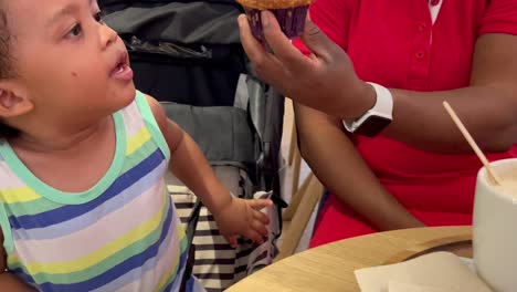 adorable y exótico niño afroeuropeo de dos años comiendo un delicioso muffin junto a su madre dentro de una cafetería