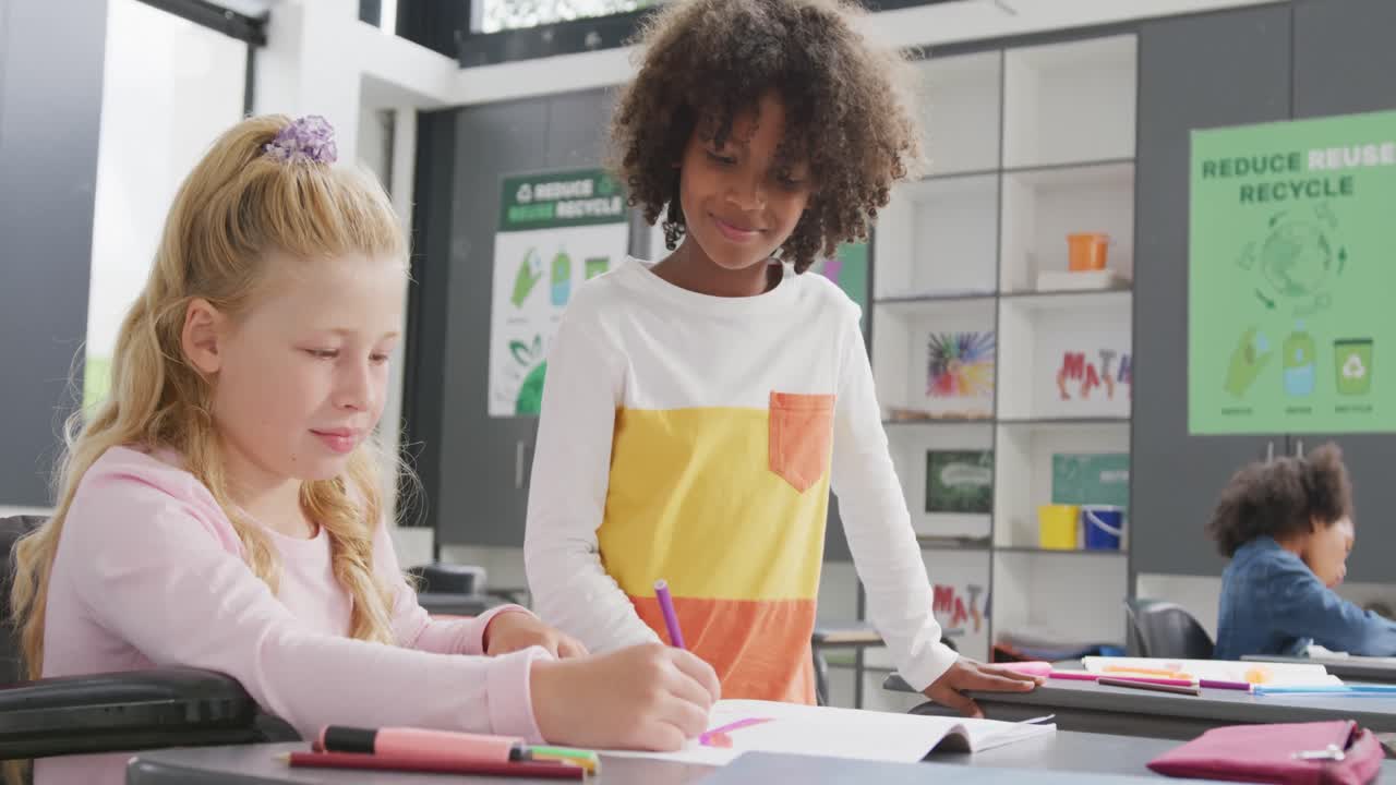 Video of happy girl in wheelchair and boy talking at desk in diverse school class