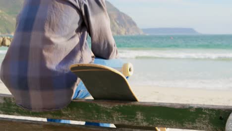 Rear-view-of-young-caucasian-man-with-skateboard-looking-at-sea-on-the-beach-4k