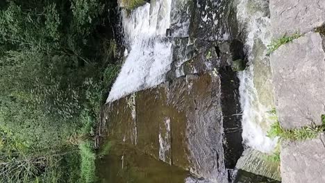 VERTICAL-slow-motion-water-cascading-down-old-Welsh-industrial-mining-stream-reservoir-towards-dense-woodland-forest